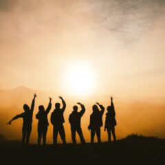 Six shadow figures arms stretched upward standing outdoors against an orange sun descending