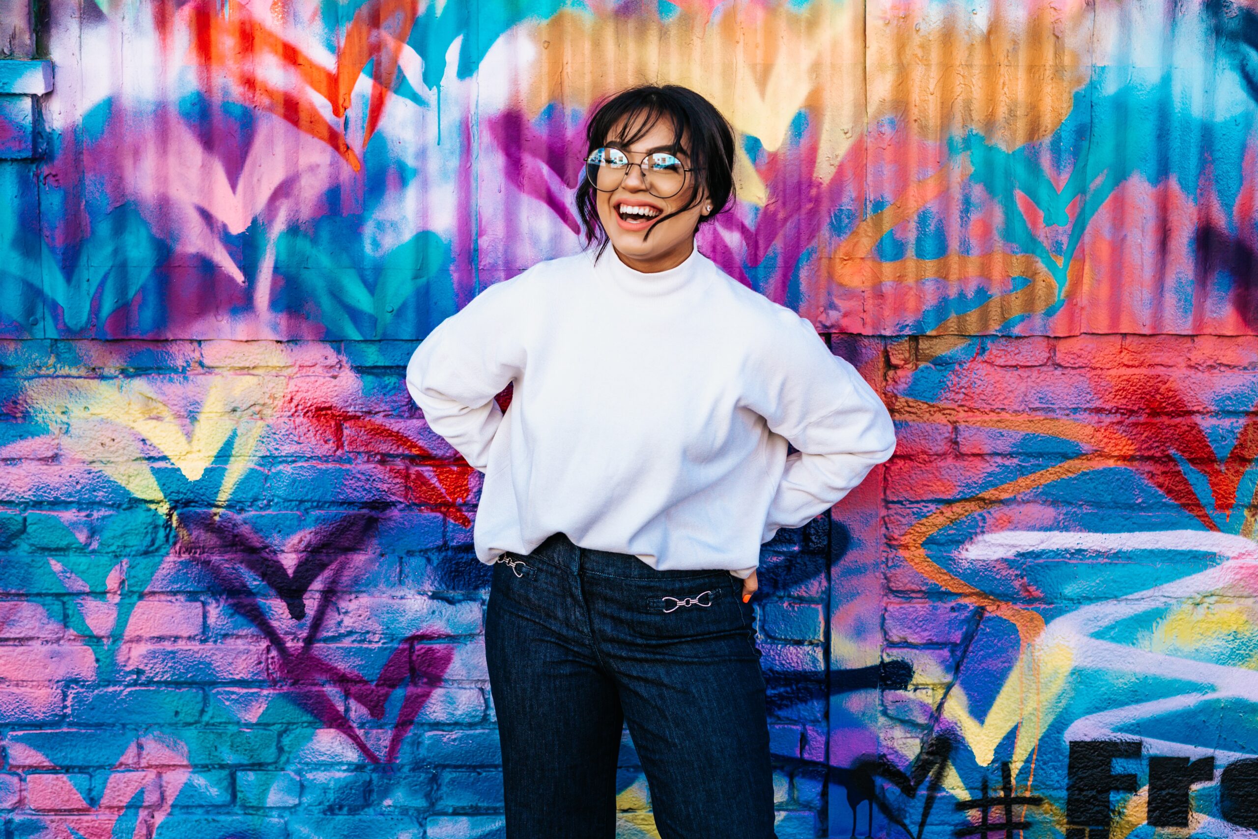Woman laughing, wearing glasses mid-thirties in dark blue pants and white fleece top, hands behing her hips, black hair pulled back stands in front of turquoise brick wall painted with graphic swanlike brush strokes of purple, black ,orange, white, rose, yellow, and green hearts