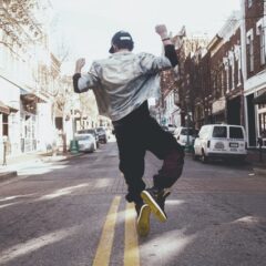 Backshot of man in black pants, black sneakers, white jacket and cap dancing in the center of the street next to two yellow parallel lineslines