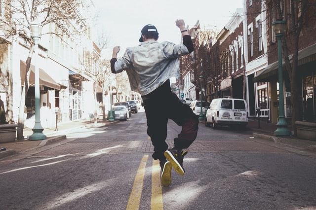 Backshot of man in black pants, black sneakers, white jacket and cap dancing in the center of the street next to two yellow parallel lineslines