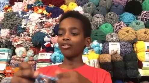 Ethiopian American boy sits and crotchets while background of colorful yarns are piled high in the background